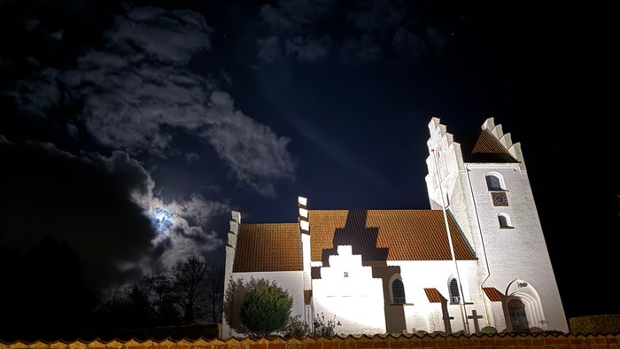 En rolig natscene med en hvid kirke, dramatiske skyer og oplyst arkitektur mod en mørk himmel.