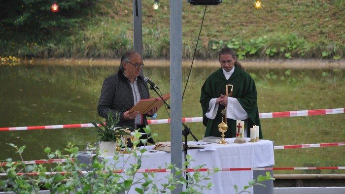 Zwei Männer stehen an einem Tisch mit Mikrofonen und Gegenständen, im Hintergrund Wasser und Bäume