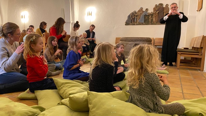 En gruppe børn og voksne sidder på gulde kuddepuder og lytter til en person i sort tøj, der står og taler.