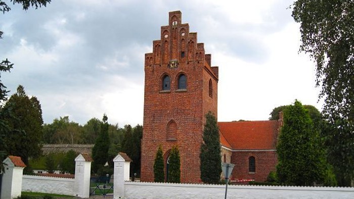 En rød murstenskirke med tårn og rødt tag, omgivet af træer og en mur.