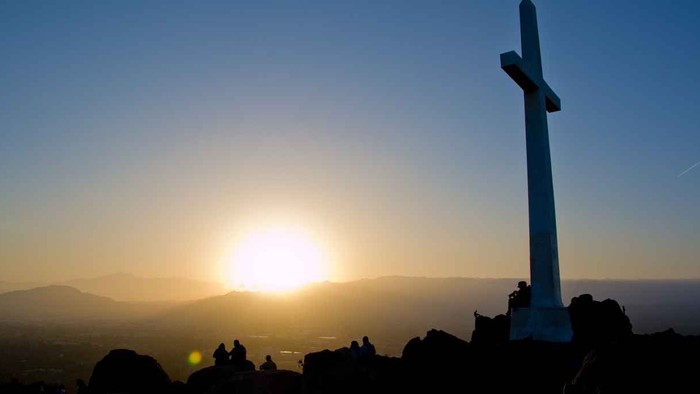 En stor korsmonument på toppen af et bjerg med solnedgang i baggrunden.