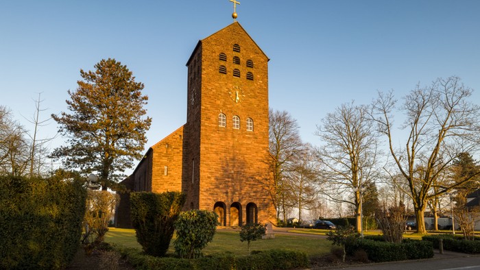 Ein Kirchengebäude aus Backstein mit hohem Turm und Kreuz, umgeben von Bäumen und Büschen.