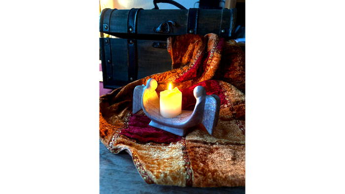 Auf einer Decke, die aus einem Koffer hängt steht ein Kerzenhalter bei dem zwei Engelsfiguren recht und links der Kerze stehen.