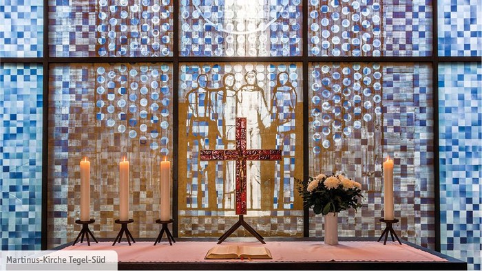 Ein Kircheninneres mit farbigem Glasfenster und einem Altar mit Kreuz und Blumen.