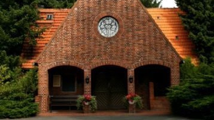 Ein hölzernes Gebäude mit Uhr und spitzem Dach, umgeben von Bäumen