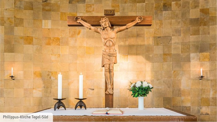 Ein Kruzifix vor einem beigefarbenen Fliesenhintergrund mit zwei Kerzen und einer Vase mit Blumen davor