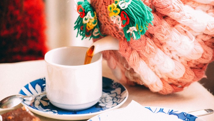 A white cup of coffee on a saucer with a spoon, next to a pink knitted object with green and yellow accents.