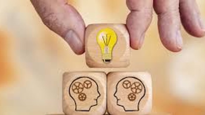 Zwei Holzwürfel mit Gehirn-Darstellungen und eine Glühbirne darüber, gehalten von einer Hand.