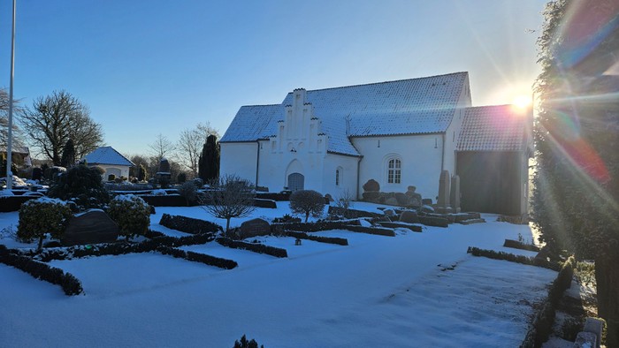 En fredfyldt vinterscene af en sneklædt kirkegård med en hvid kirke i baggrunden under klart solskin.