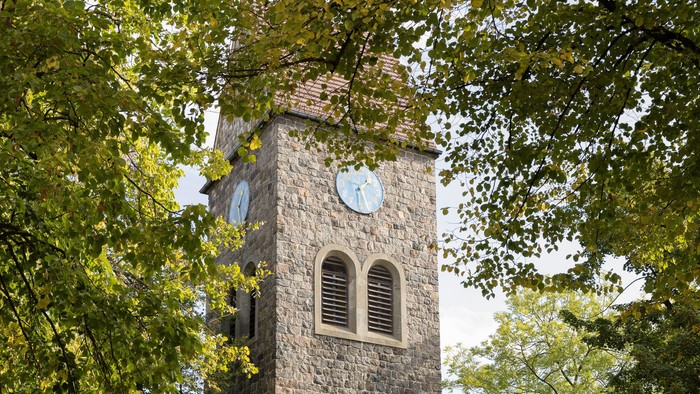 Der Kirchturm aus Stein mit Uhr und Fenster steht in einem Park.