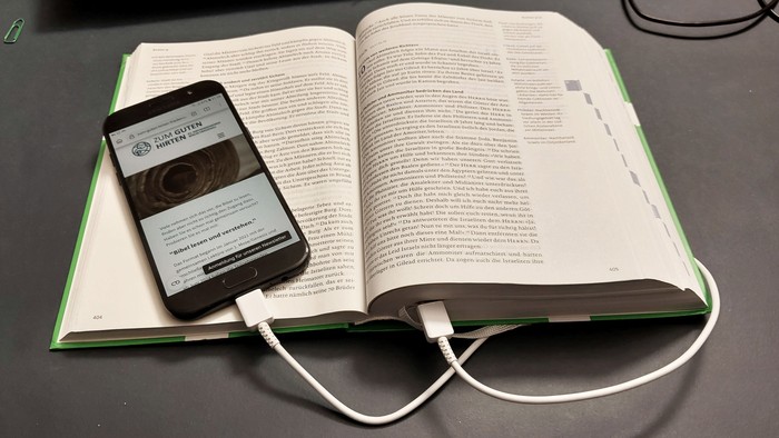 Ein Smartphone liegt auf einem aufgeschlagenen Buch, verbunden mit einem Ladekabel