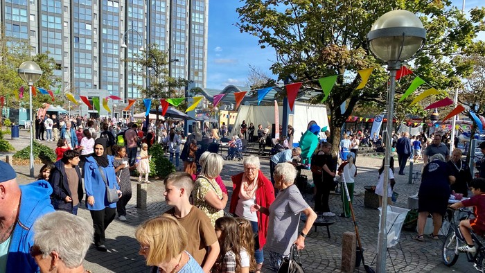 En folkemængde på en plads med farverige flag og høje bygninger i baggrunden