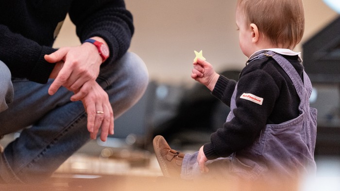 Ein Baby sitzt auf dem Boden und hält ein gelbes Spielzeug, während eine erwachsene Person daneben kniet.