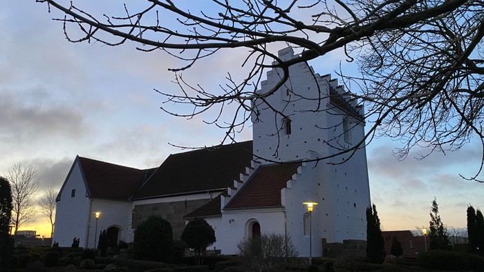 En hvid kirke med tårn og røde tag ved solnedgang, omgivet af træer og buske