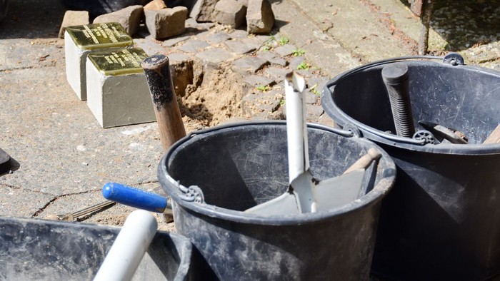 Kurz vor einer Verlegung von Stolpersteinen im Straßenpflaster: Werkzeug im Vordergrund, im Hintergrund zwei Stolpersteine