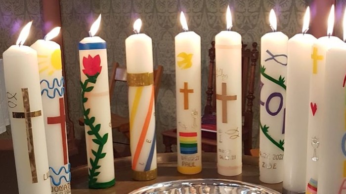 Ein runder Tisch trägt brennende Kerzen mit verschiedenen Symbolen und ein zentrales silbernes Becken in einer geschmückten Kapelle.