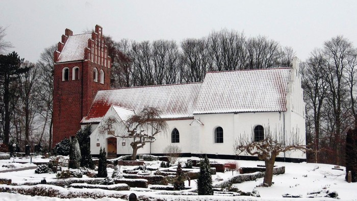 En snedækket kirke med et rødt tårn og et hvidt tag, omgivet af bærbare træer og sne.
