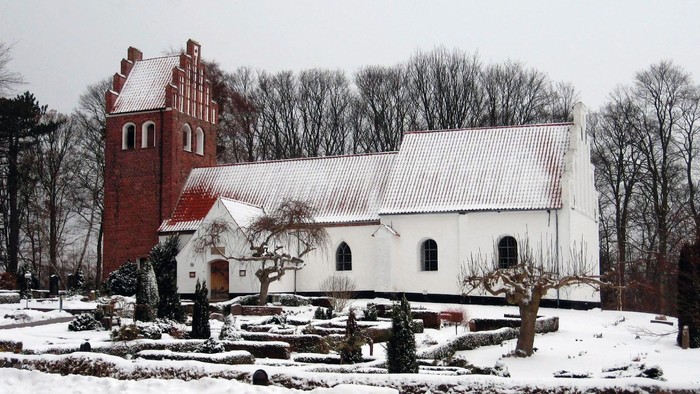 En snedækket kirke med et rødt tårn og et hvidt tag, omgivet af sne og træer