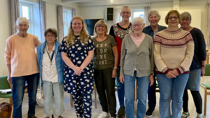 En gruppe ældre og en yngre kvinde står sammen i et rum.