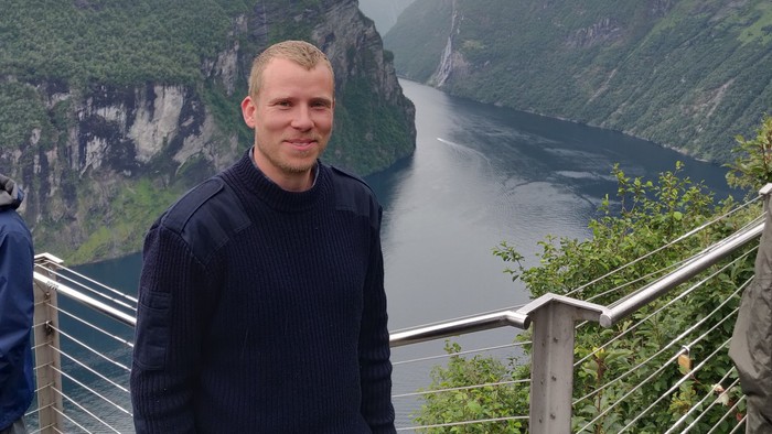 En person står og smiler ved en naturskøn fjord omgivet af stejle, frodige grønne klipper.