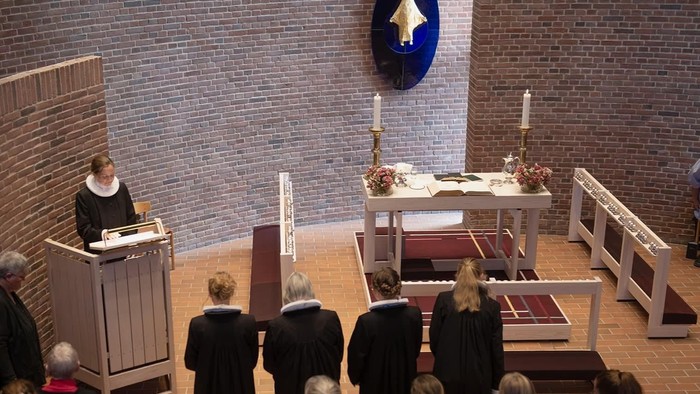 Kirketjeneste med præst ved prædikestolen, alter med stearinlys og menighed.