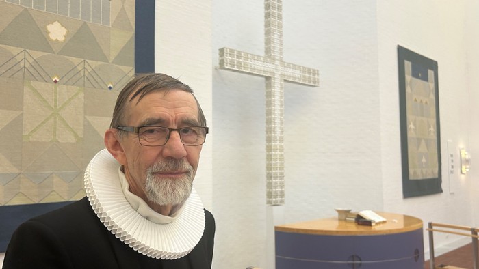 En mand i gejstligt tøj står ved siden af et stort kors i en pyntet indendørs omgivelse.