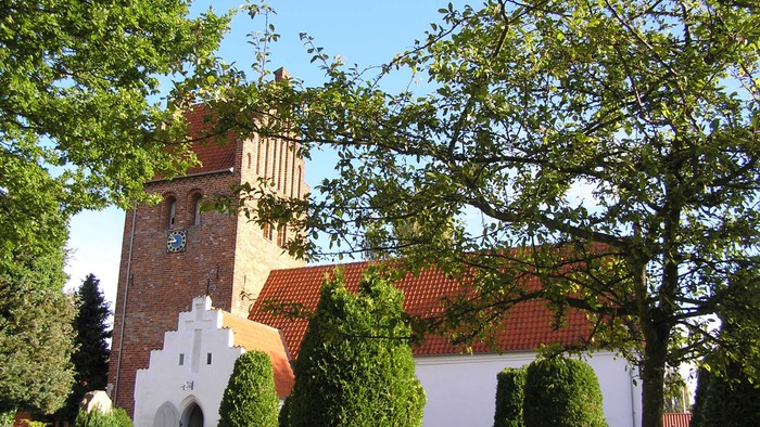 Et hvidt kirkebygning med røde tag og en tårn, omgivet af grønne træer og buske