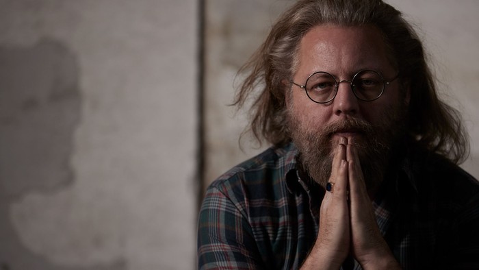 En langhåret mand med briller og skæg sidder med håndene samlet foran munnen.