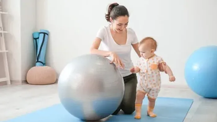 Eine Frau hilft einem Baby, auf einem Gymnastikball in einem Fitnessraum zu stehen.