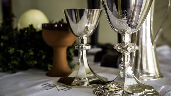 Drei silberne Kelche auf einer weißen Tischdecke mit Grünpflanzen im Hintergrund.