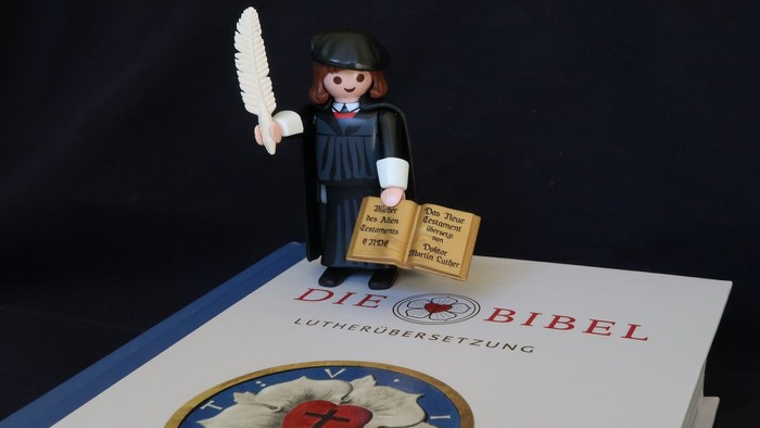 Eine Playmobil-Figur hält ein Buch und eine Feder über einem Buch mit dem Titel