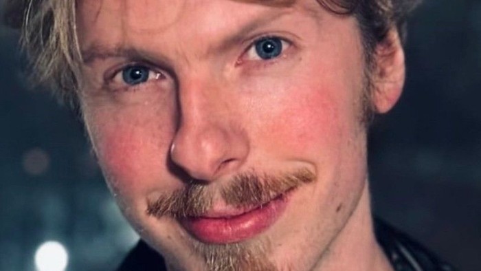 En mand med blondt hår og skæg, iført en mønstret jakke og et sølvkædehalsbånd, smiler til kameraet.