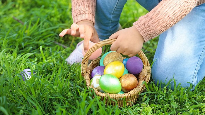 Ein Kind hält einen Korb mit bunt bemalten Ostereiern auf einem grasbewachsenen Bereich.
