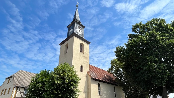 Kirchturm mit Uhr und Kreuz, umgeben von Bäumen und Häusern