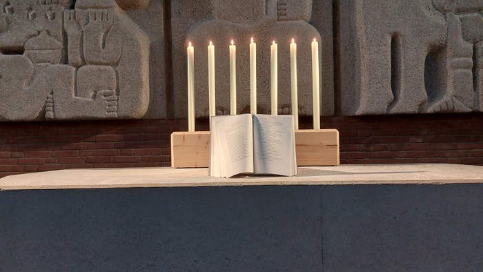 Offenes Buch mit brennenden Kerzen darauf, vor einer strukturierten Wand.