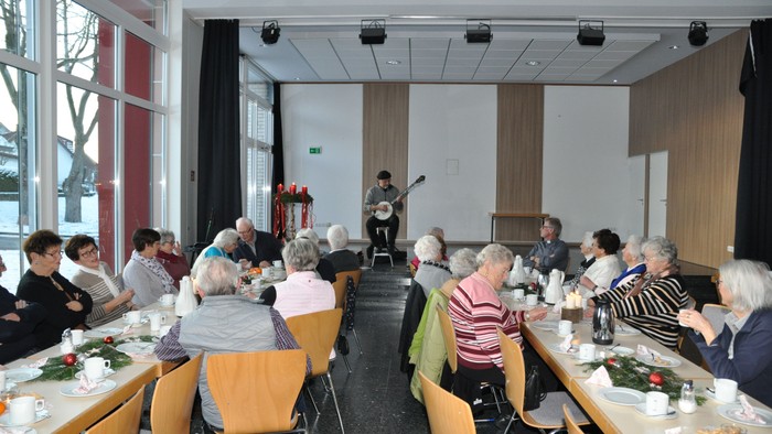 Eine Gruppe älterer Menschen sitzt an Tischen und hört einem Mann zu, der vorne steht und spricht.