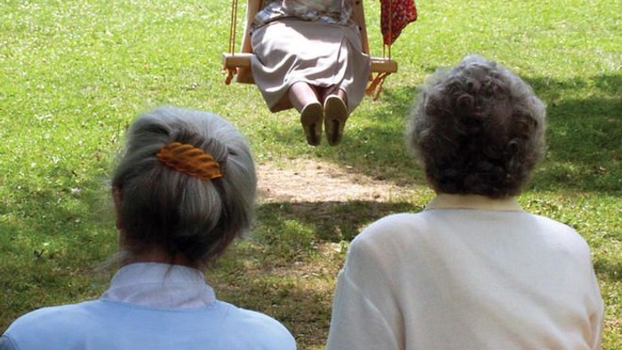 Alte Frau schaukelt im Park, zwei Frauen sitzen auf Bank und schauen zu