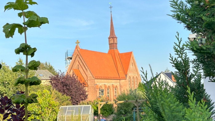 Ein rotes Kirche mit spitzem Turm, umgeben von grünem Gebüsch und parkenden Autos.