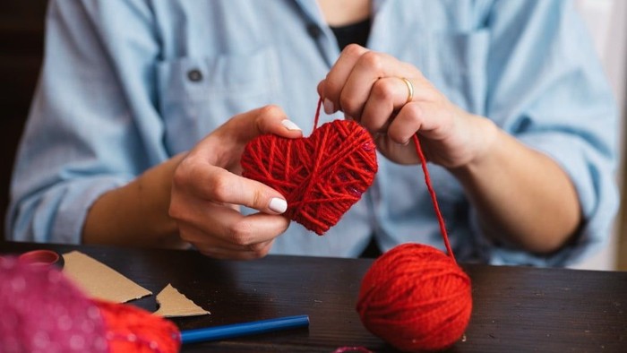 En person sitter ved et bord og laver et hjerte af røde tråder.