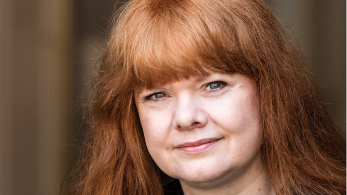 En person med langt rødt hår iført en sort top poser mod en neutral baggrund.