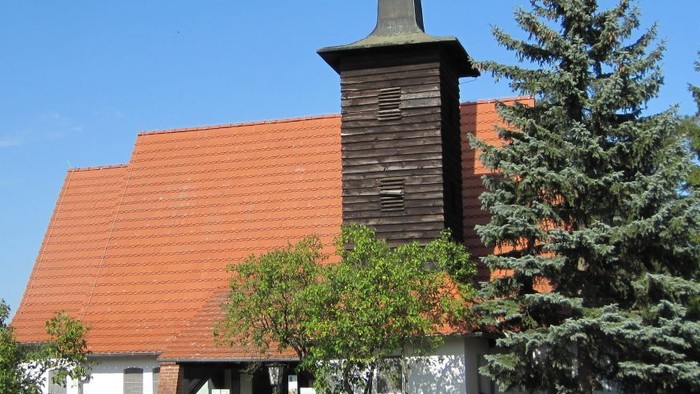 Kirchengebäude mit hohem Turm und rotem Dach, umgeben von Bäumen und Büschen