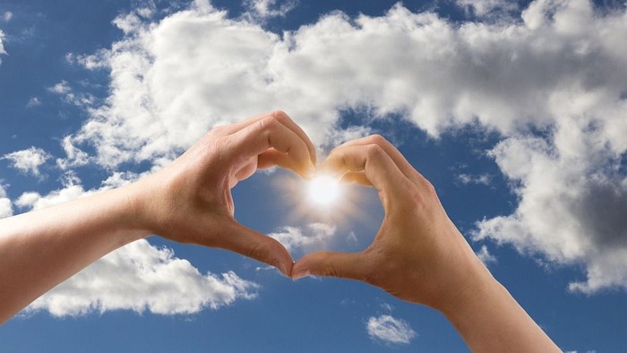 Zwei Hände bilden ein Herz vor einem sonnigen Himmel