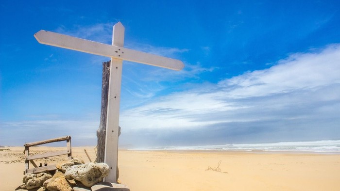 Ein hölzernes Kreuz steht auf einem Steinhaufen am sandigen Strand unter einem klaren blauen Himmel.