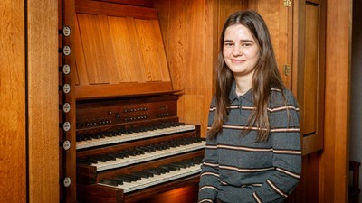 En ung person smiler ved siden af et stort træpibeorgel med flere sæt tangenter.