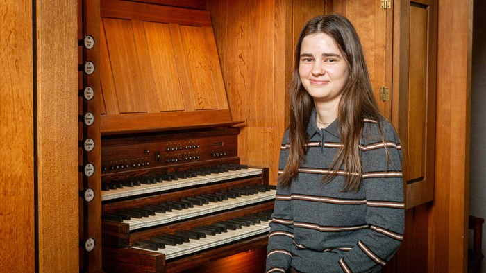 En ung person smiler ved siden af et stort træpibeorgel med flere sæt tangenter.