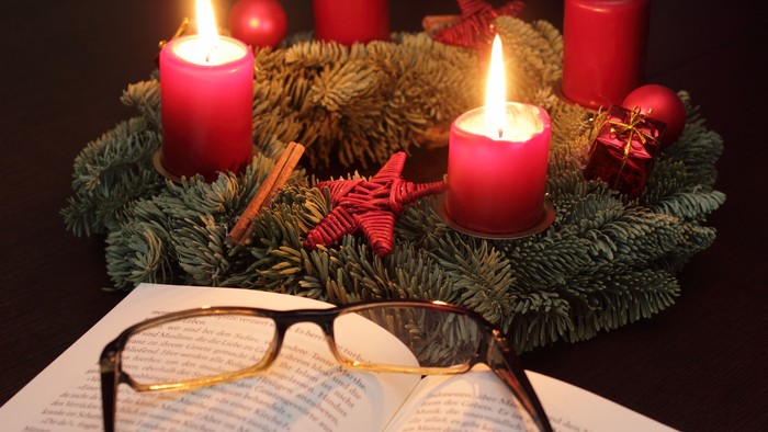 Im Vordergrund ein Buch mit darauf liegender Brille, im Hintergrund ein Adventskranz mit vier roten Kerzen, von denen zwei brennen