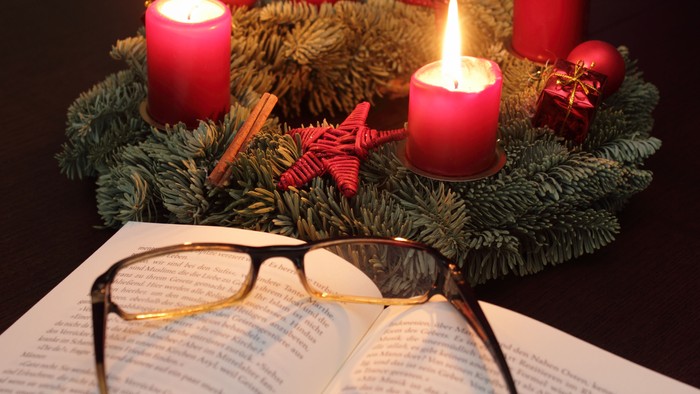 Im Vordergrund ein Buch mit darauf liegender Brille, im Hintergrund ein Adventskranz mit vier roten Kerzen, von denen zwei brennen