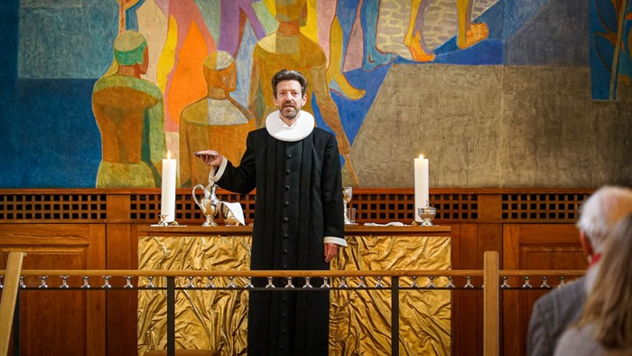 En mand i svart kapittel holder en lille hund i en kirke.