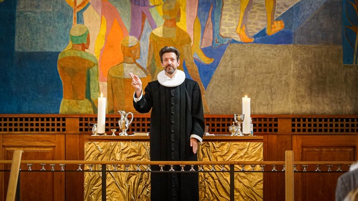 En præst i svart kappe og halsbånd holder en hostia op forud for alteret i en kirke.