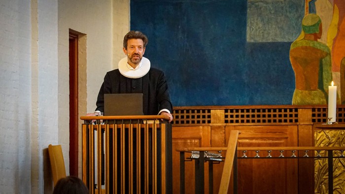 En mand i mørkt tøj og halsbånd står ved et podium og holder en laptop.